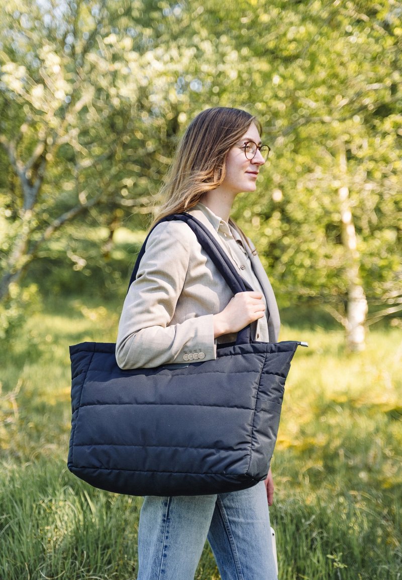 Svart puffig handväska med två stadiga axelremmar, rektangulär form och sydd textur, buren av en person i en naturlig miljö.