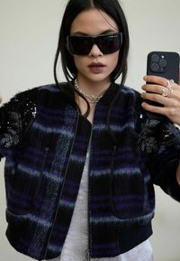 Czarna i fioletowa kraciasta kurtka bomberka z teksturowanej tkaniny, kieszeniami na zamek, koronkowymi akcentami na ramionach oraz ściągaczem u dołu.