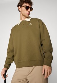 Olivengrønn genser med en kremfarget krage, med et lite brodert logo. Stoffet ser mykt ut med ribbede mansjetter og nederkant.