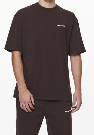 Mørkebrun bomulds T-shirt med korte ærmer, med et hvidt "PEGADOR" logo på venstre bryst. Løs pasform, glat tekstur.