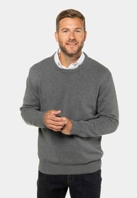 Grauer Strickpullover mit rundem Halsausschnitt und gerippten Bündchen. Kombiniert mit dunklen Jeans, die Hände sind zusammen in einem neutralen Hintergrund gezeigt.