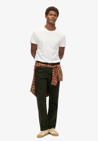 Camiseta blanca de algodón, pantalones verde oscuro, camisa a cuadros naranja anudada a la cintura, zapatos beiges con cordones blancos, de pie sobre un fondo blanco.