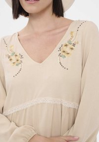 Beige jurk met een V-halslijn, bloemige borduursels in geel en groen, kanten rand bij de taille enGathered mouwen voor een vloeiende silhouet.