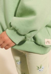 Sweatshirt vert menthe avec des poignets côtelés, associé à un legging vert clair ornés de motifs floraux. Texture subtile et détail de logo visibles.