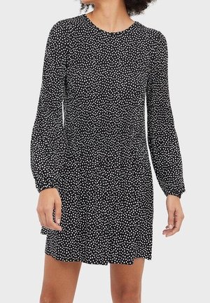 Frau trägt ein schwarzes Langarmkleid mit weißen Punkten, elastischer Taille und einem Faltenrock, stehend vor einem schlichten Hintergrund.