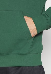 Main avec un tatouage "TIME" sur le poignet à l'intérieur de la poche d'un sweat à capuche vert porté avec un pantalon noir.