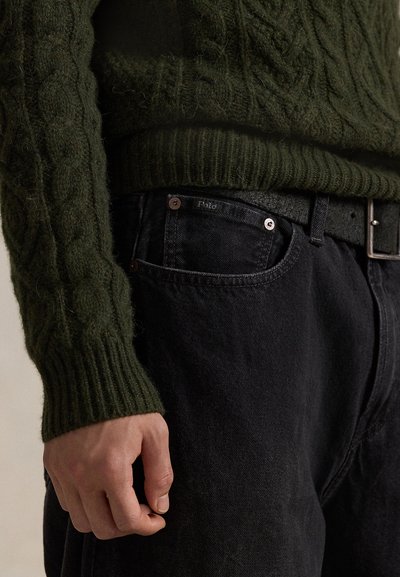 Hånd hvilende ved lommen på svarte jeans med "Polo" merke, iført en mørkegrønn kabelstrikket genser stukket inn i jeans med svart belte.