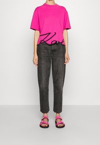 Rosa Baumwoll-T-Shirt mit schwarzem, geschwungenem "Karl"-Druck, kombiniert mit dunkelgrauen Jeans und Sandalen mit rosa Riemen. Lässige Outfit-Präsentation.