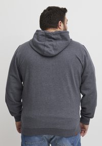 Felpa grigia con cappuccio dal tocco morbido, dotata di tasca a marsupio, polsini e orlo a coste, e un'etichetta con logo in basso a sinistra.