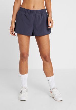 Kobieta ubrana w granatowe sportowe szorty, białe skarpetki Nike i białe tenisówki, stojąca na jasnym, jednolitym tle.