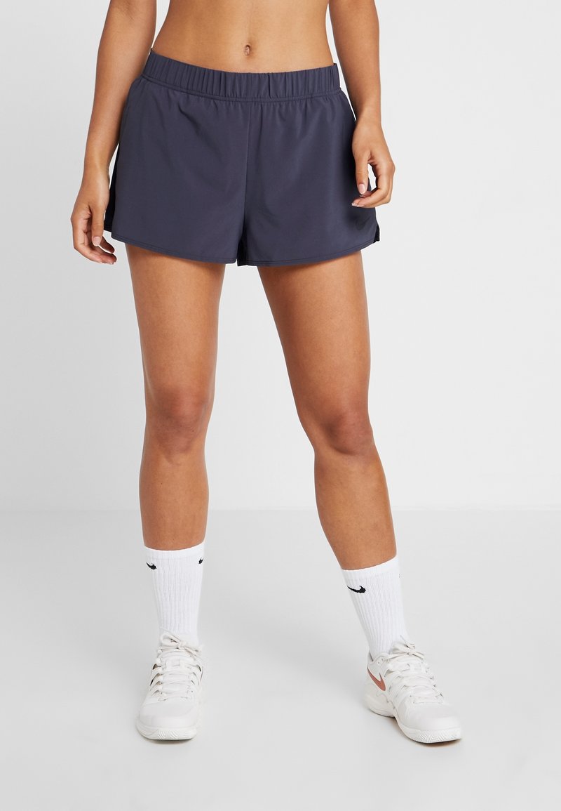 Femme portant un short de sport bleu marine, des chaussettes blanches Nike courtes et des baskets blanches, debout devant un fond uni clair.