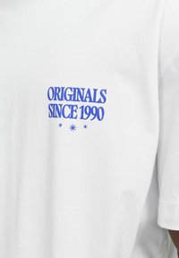 Camiseta de algodón blanca con texto bordado en azul "ORIGINALS SINCE 1990" con acentos de estrellas, mangas cortas y cuello redondo.