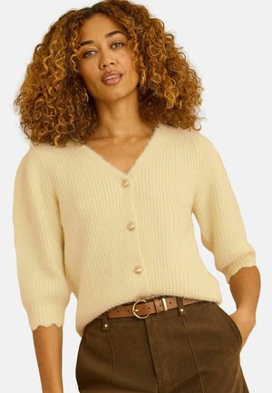 Käärus juustega naine, kes kannab koorevärvi soonikkoes nööpidega kardigani ja pruune kõrge vöökohaga pükse, millel on beež vöö, poseerides ühe käega taskus.