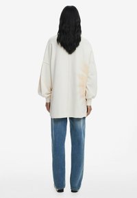 Felpa oversized color crema con spalle drop, dal texture morbido e con spacchi laterali, abbinata a jeans blu larghi e scarpe nere.