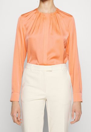 Mujer que lleva una blusa de satén melocotón con mangas largas y escote fruncido, metida dentro de pantalones de talle alto a medida de color blanco roto.