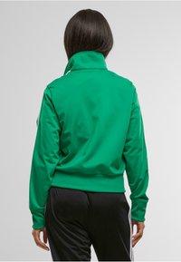 Chaqueta deportiva verde con un alto cuello y mangas largas, que presenta una textura suave. Pantalones negros complementan el conjunto.