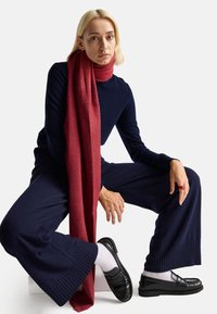 Sciarpa di lana rossa drappeggiata sulle spalle; modella che indossa un top di maglia scuro e pantaloni navy a gamba larga; presenta mocassini neri con calze bianche.