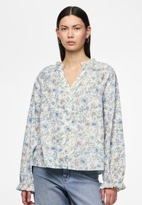 Blouse légère à boutons, avec un motif floral bleu et pêche, col froncé et manches longues, associée à un jean en denim.