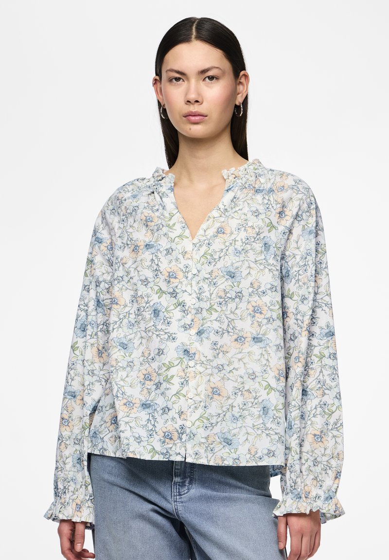 Blouse légère à boutons, avec un motif floral bleu et pêche, col froncé et manches longues, associée à un jean en denim.