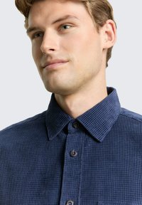 Man met lichte huid en blauwe ogen, gekleed in een donkermblauwe gestructureerde overhemden met knopen, kijkt iets opzij tegen een neutrale achtergrond.