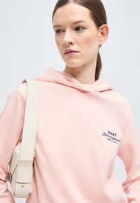 Ženska svetle kože z rjavimi lasmi, zvezanimi v čop, oblečena v bledo roza kapucar in nosi bež torbico čez ramo, gleda levo.