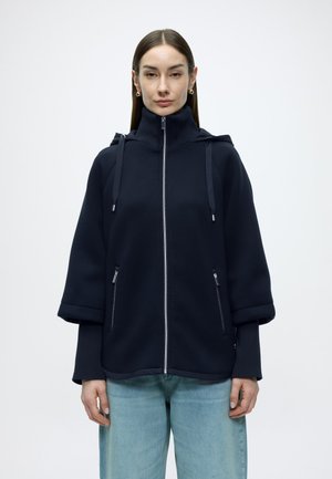 Navy zip-up jacket with a high collar, large hood, and ribbed cuffs. Features two zippered side pockets and a relaxed fit.