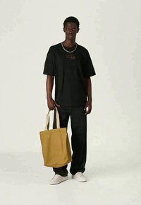 Tricou negru supradimensionat cu un logo, combinat cu pantaloni negri cu croială lejeră și adidași albi. Ține o geantă de umăr bej cu bretele deschise.