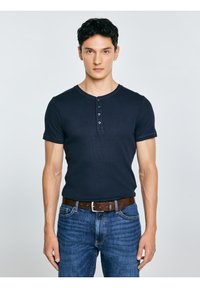 Joven con cabello corto y oscuro, vestido con una camisa Henley de manga corta azul marino, jeans azules y un cinturón de cuero marrón, de pie frente a un fondo claro.