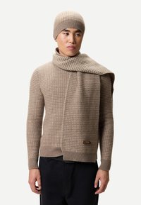 Beige stickad tröja med ett strukturerat mönster, matchande halsduk hängd över. Den stickade mössan kompletterar setet. Tröjan har en logotypetikett.