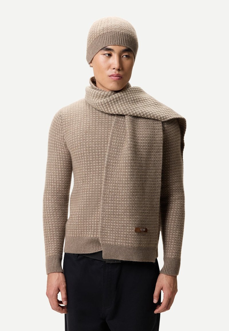 Beige stickad tröja med ett strukturerat mönster, matchande halsduk hängd över. Den stickade mössan kompletterar setet. Tröjan har en logotypetikett.