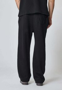 Homme portant un pantalon noir ample avec une poche arrière et des chaussures sombres, se tenant devant un fond léger gris uni.