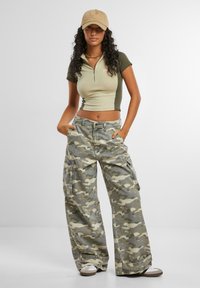 Kuttet olivengrønn zip-up genser med korte ermer; grå camo cargo bukser med lommer. Nøytral baseballcap; hvite og brune sneakers.
