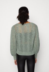 Pull vert menthe en tricot avec un motif en dentelle, ourlet côtelé et manches bouffantes. Porté avec des leggings noirs, vu de dos.