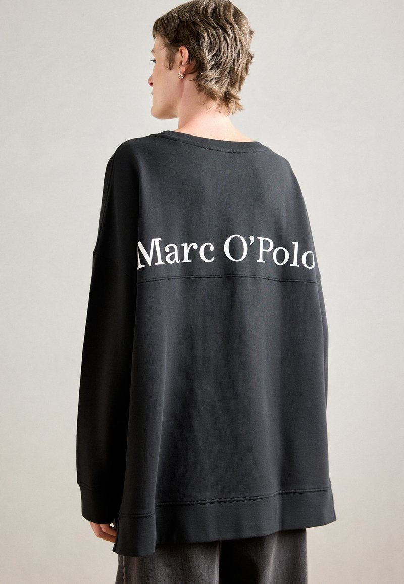 Črna oversized pulover z mehkim materialom, s rebrastimi manšetami in izrazitim belim logotipom "Marc O'Polo" na hrbtni strani.