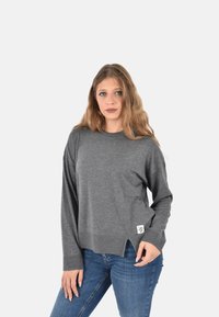 Giovane donna con capelli lunghi che indossa un maglione grigio e jeans blu, in piedi contro uno sfondo chiaro e uniforme.