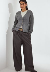 Grau gestrickte Strickjacke mit V-Ausschnitt, Knopfverschluss und Taschen, kombiniert mit weiten dunkelgrauen Hosen und einem beigen Oberteil.