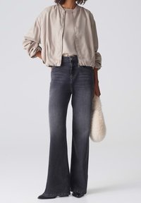 Chaqueta bomber beige claro con textura suave, vaqueros acampanados de talle alto en gris y un bolso de mano de piel sintética en color crema.