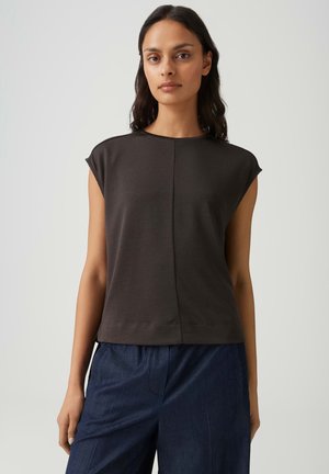 Femme aux cheveux foncés portant un haut noir sans manches et un pantalon bleu foncé, debout devant un fond clair uni.