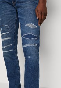 Jean bleu avec un design déchiré comportant plusieurs zones rippées et des bords effilochés. Style classique à cinq poches avec une coupe droite.