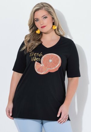 Lyshåret kvinde iført sort skjorte med teksten "Fresh Vibes" og en grafik af en appelsinskive, gule blomsterøreringe og lyseblå bukser.