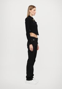 Black cropped jacket with a collar and gold accents, paired with black pants and white sneakers. Smooth fabric, fitted design, side view.