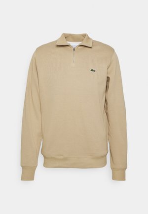 Pull-over beige à manches longues avec un col zippé. Il présente une texture côtelée et un petit logo vert Lacoste sur la poitrine gauche.
