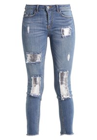 Vaqueros azules desgastados con cinco desgarros prominentes, que revelan parches de malla plateada. Diseño ajustado con dobladillo deshilachado y textura clásica de denim.