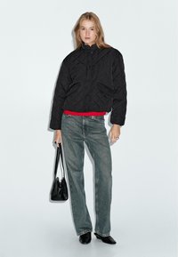 Giacca nera trapuntata con colletto alto, cintura rossa, abbinata a jeans blu chiaro a gamba dritta e borsa nera. Il modello indossa scarpe nere.
