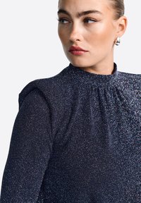 Blusa azul marino con un escote alto, detalles de fruncido en los hombros y tela texturizada con sutiles destellos iridiscentes.