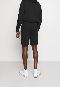 Schwarzer Kapuzenpullover kombiniert mit schwarzen Shorts, die einen lässigen Schnitt, Taschen und weiße High-Top-Sneaker aufweisen. Keine sichtbaren Logos oder Muster.