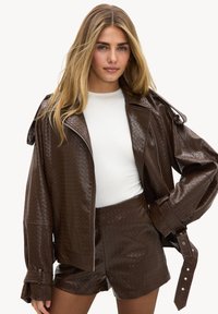 Femme aux longs cheveux blonds portant une veste en cuir brun texturé et un short assorti par-dessus une chemise blanche, posant avec une main sur la hanche.