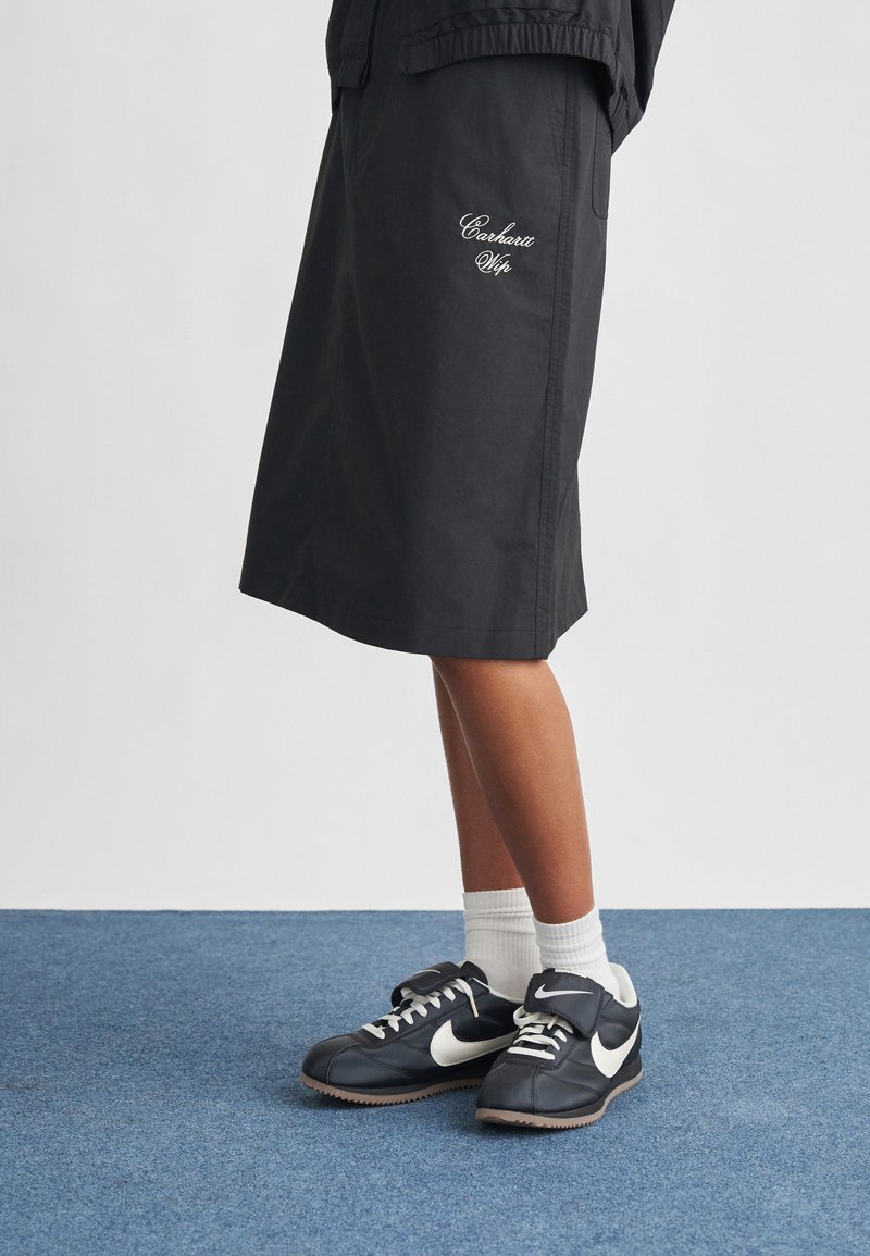 Black knee-length skirt made of lightweight material, featuring white "Carhartt WIP" embroidery. Paired with black Nike sneakers and white socks.
