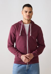 Maroon zip-up dukserica s kapuljačom, prednjim džepovima i rebrastim manžetama. Nošena preko bijele majice, na neutralnoj pozadini.