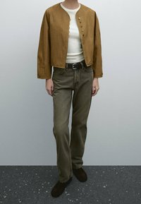 Giacca marrone con scollatura rotonda e chiusura a bottoni, indossata sopra un top crema e jeans marrone-verde. Scarpe ankle boots marroni completano il look.